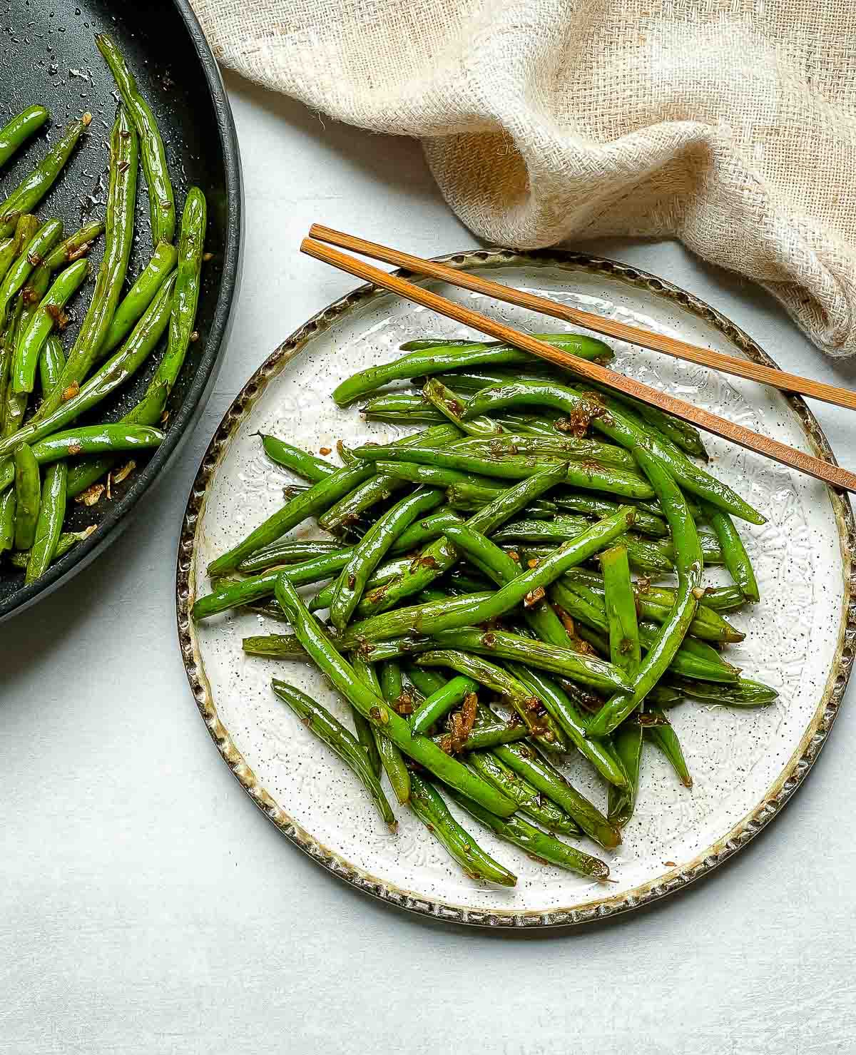Chinese Garlic Green Beans on a round white plate with a pair of brown chopsticks and green beans in a frying pan on the side with a napkin.