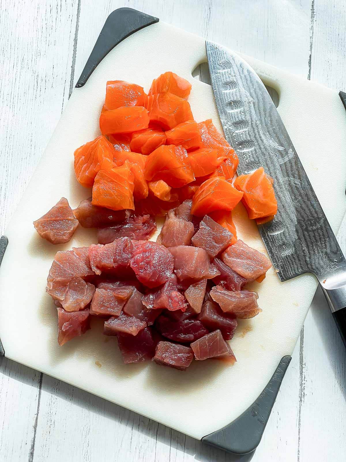 Cubed salmon and tuna on a white cutting board with a chef's knife on the side.