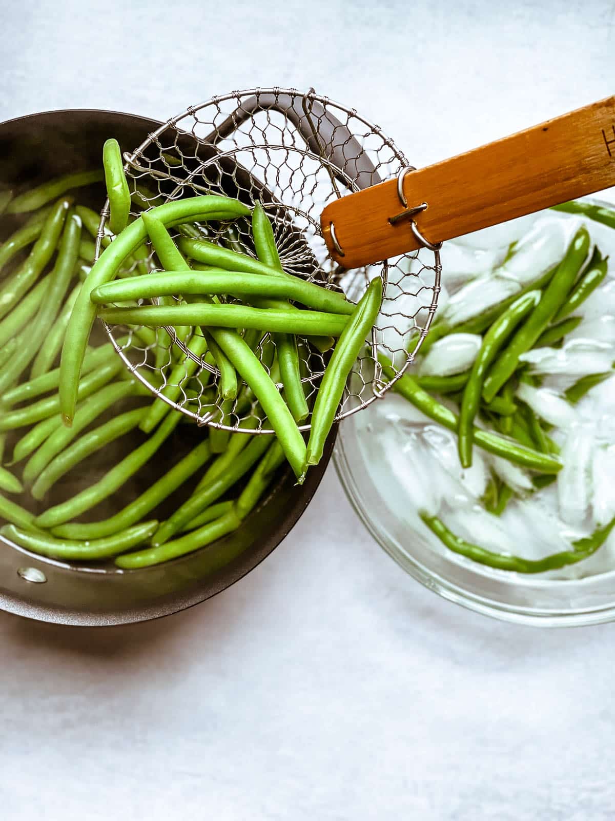 A spider strainer lifting green beans out of a pot of cooked green beans and green beans in a bowl of ice water on the side.