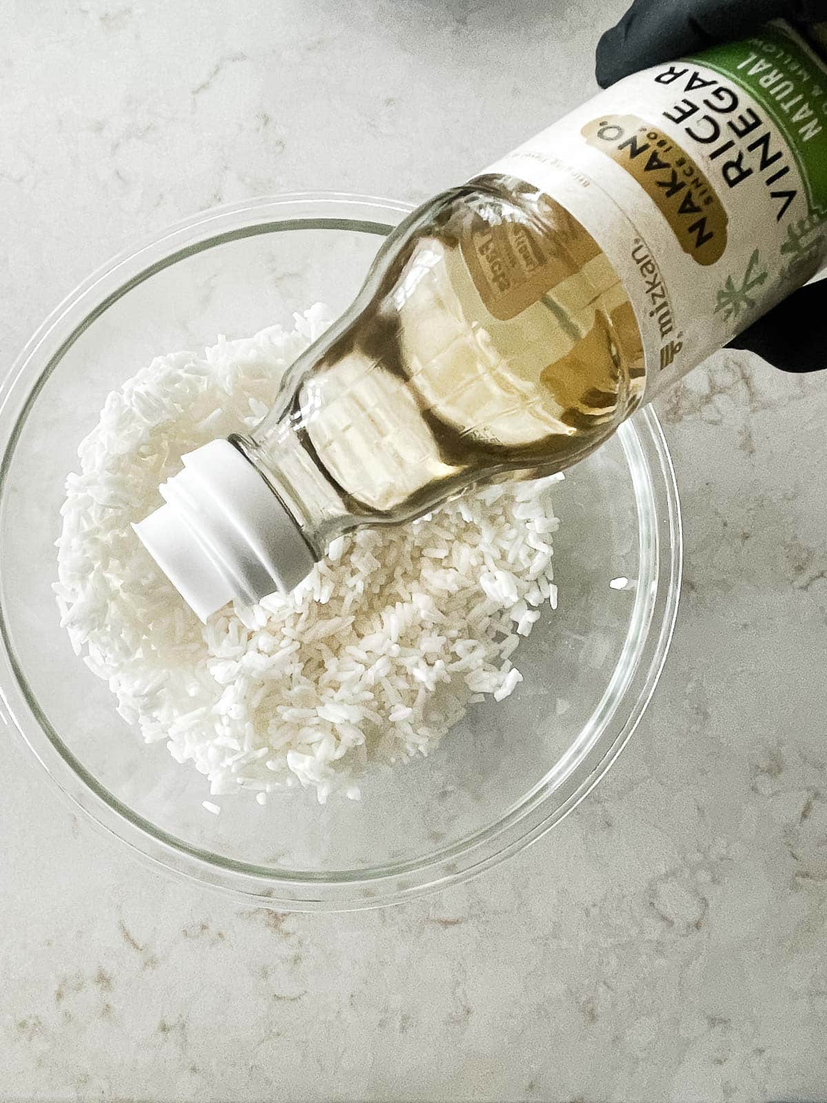 Rice vinegar being poured into cooked white rice in a glass bowl.