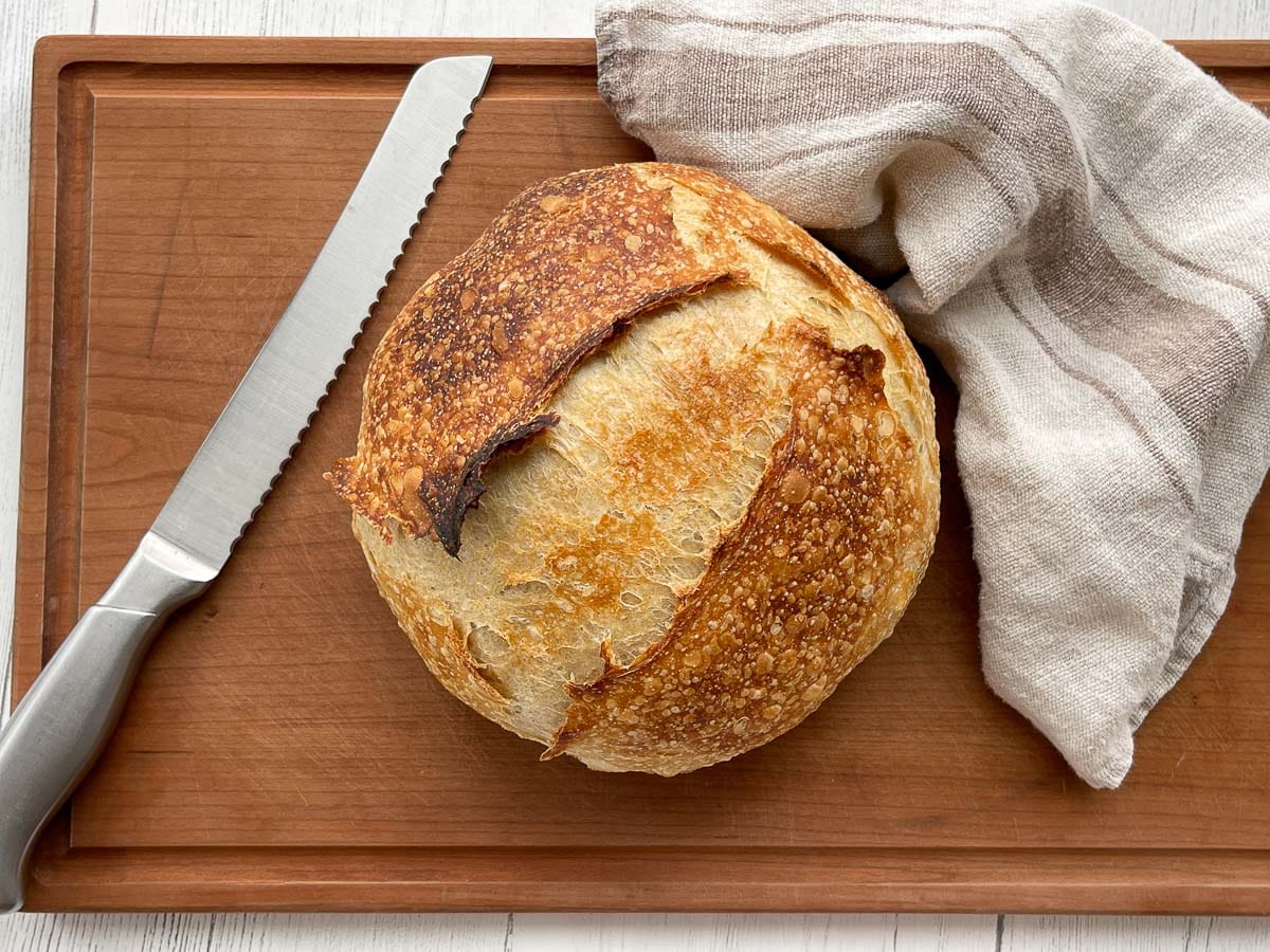 A round sourdough bread on top of a wooden cutting board with a bread knife and linen napkin on the side.