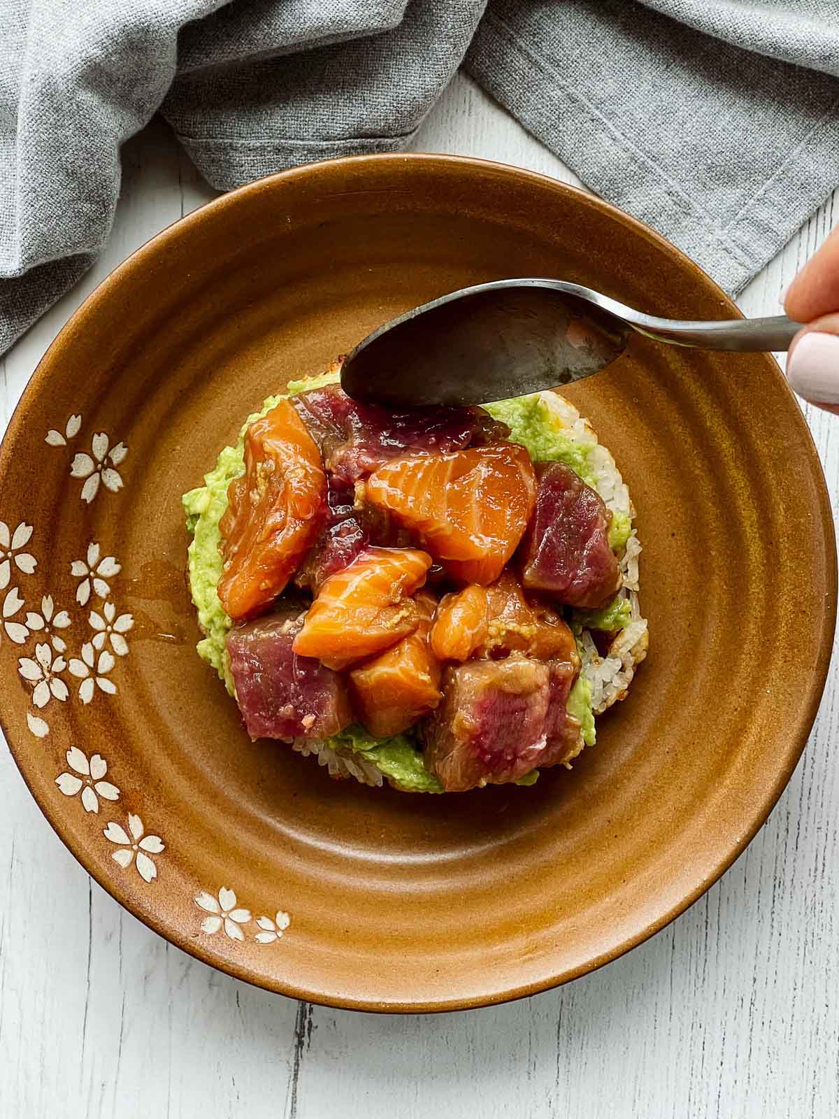 A hand spooning cubed salmon and tuna on top of a sushi waffle on a brown plate.
