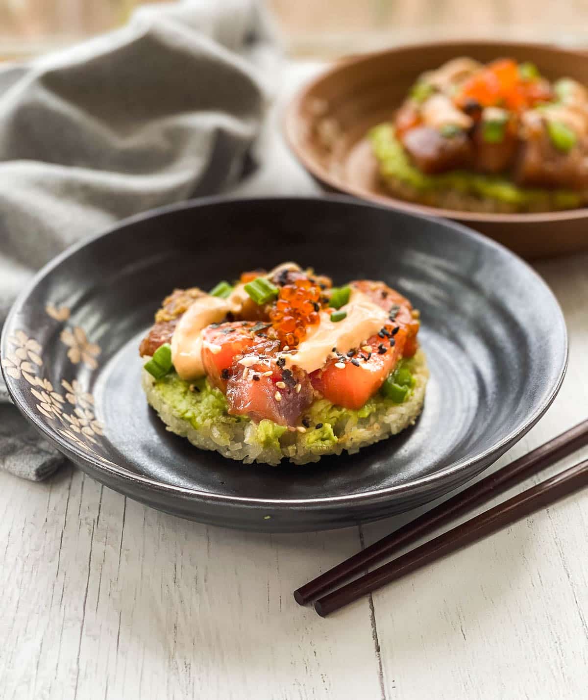 Individual sushi waffles on round plates featuring cubed sushi-grade salmon and tuna with a side of chopsticks on top of a white surface with a napkin in the background.