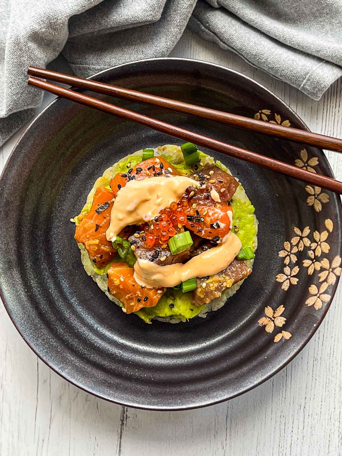 A sushi waffle with salmon and tuna on a round black plate with chopsticks on top of a white surface with a napkin on the side.