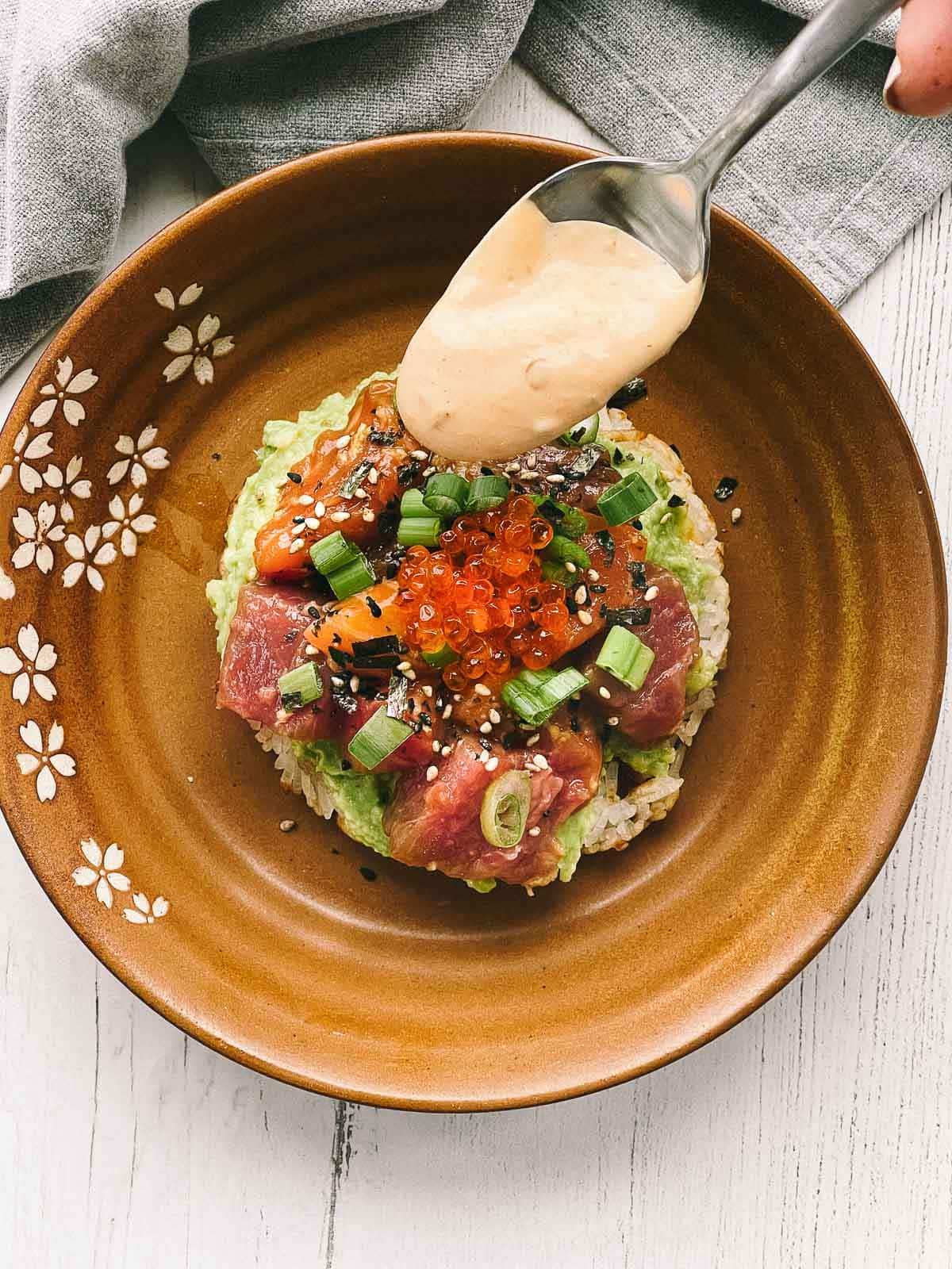 A spoon with aioli being drizzled on top of a sushi waffle on a brown plate.