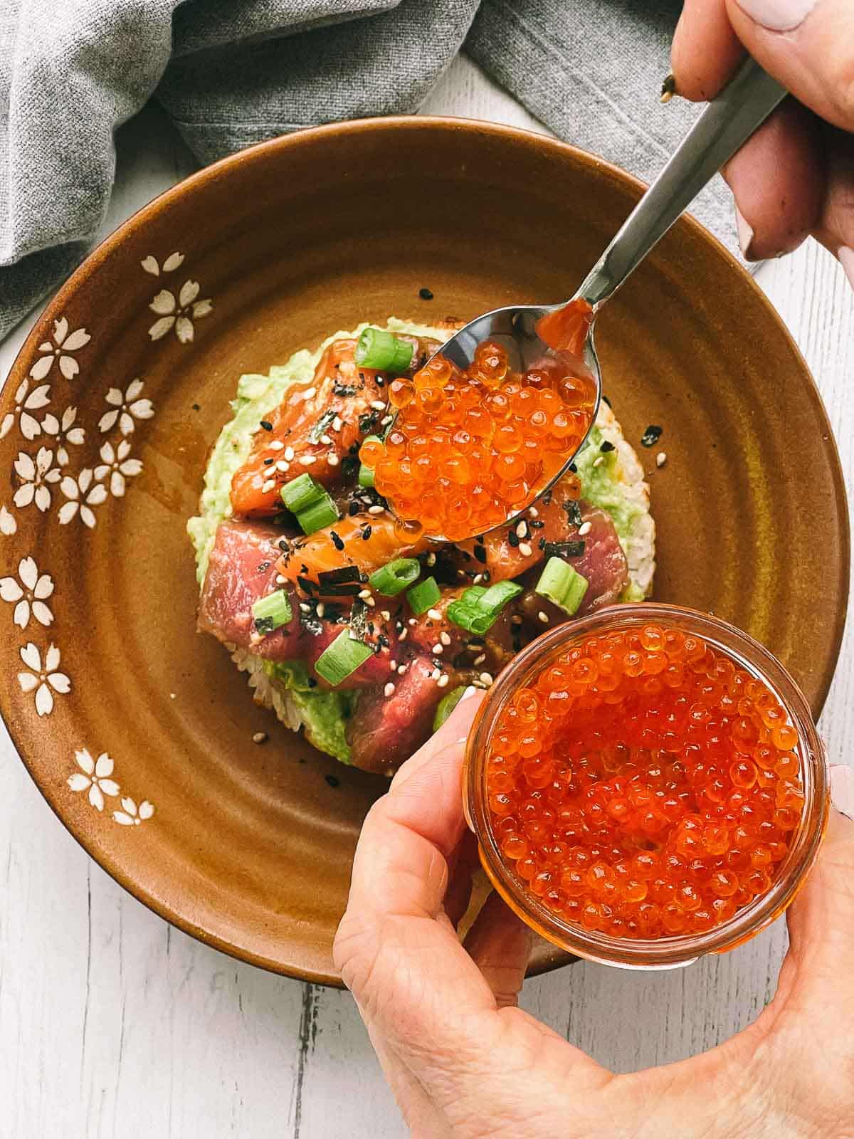 A spoon with salmon roe caviar being placed on top of a sushi waffle on a brown plate.