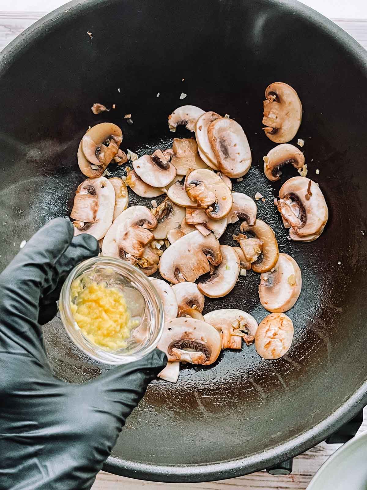 A hand adding grated ginger in a small glass bowl to stir-fried button mushroom slices in a hot wok.