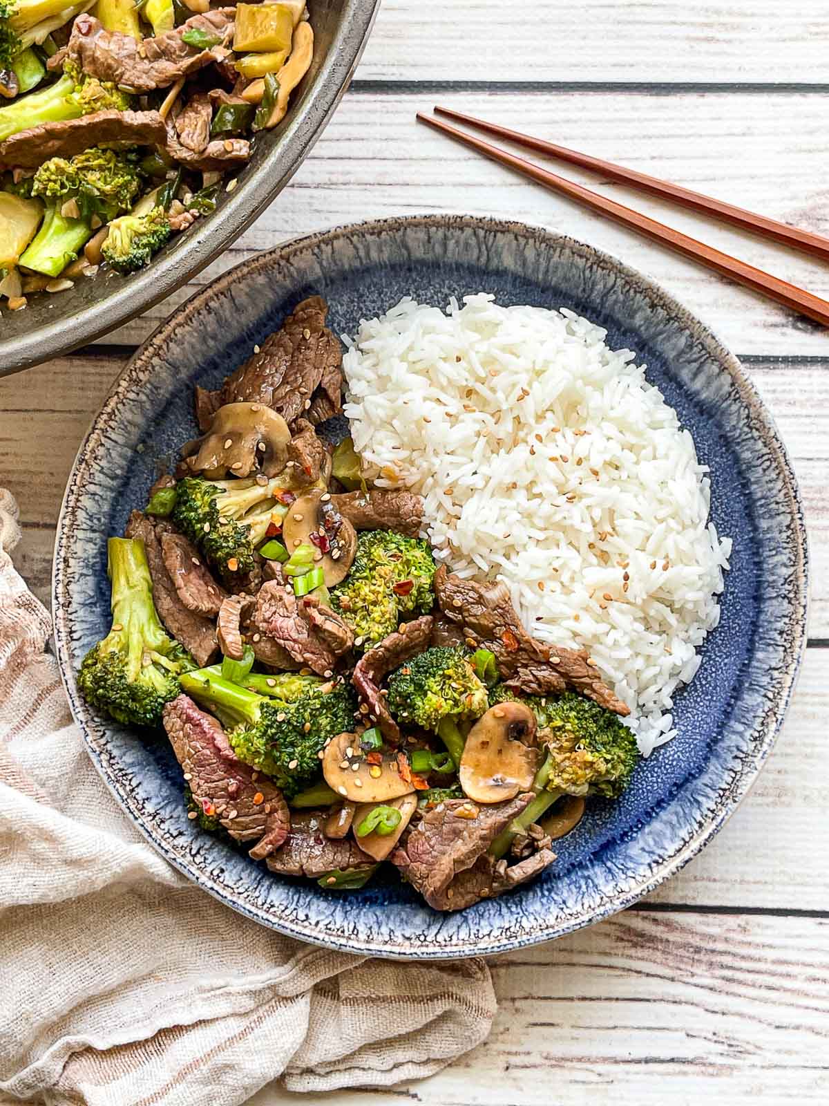 Beef and broccoli stir fry and white rice in a round blue bowl and a wok filled with stir fry, with a pair of wooden chopsticks and a linen napkin on the side, on top of a white wooden board.