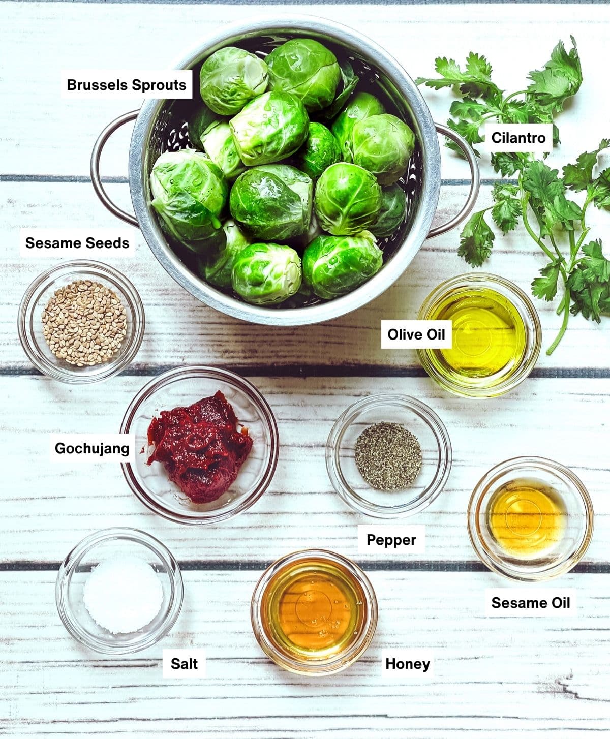 Brussels sprouts in a silver bowl and gochujang sauce ingredients in small glass bowls on top of a white wooden board.