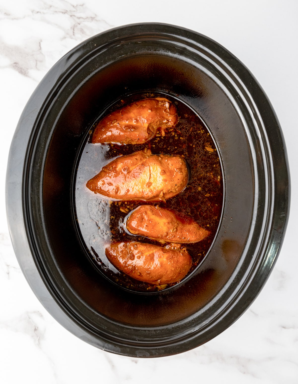 Four cooked chicken breast fillets bathing in a honey garlic sauce, on top of a marble surface.