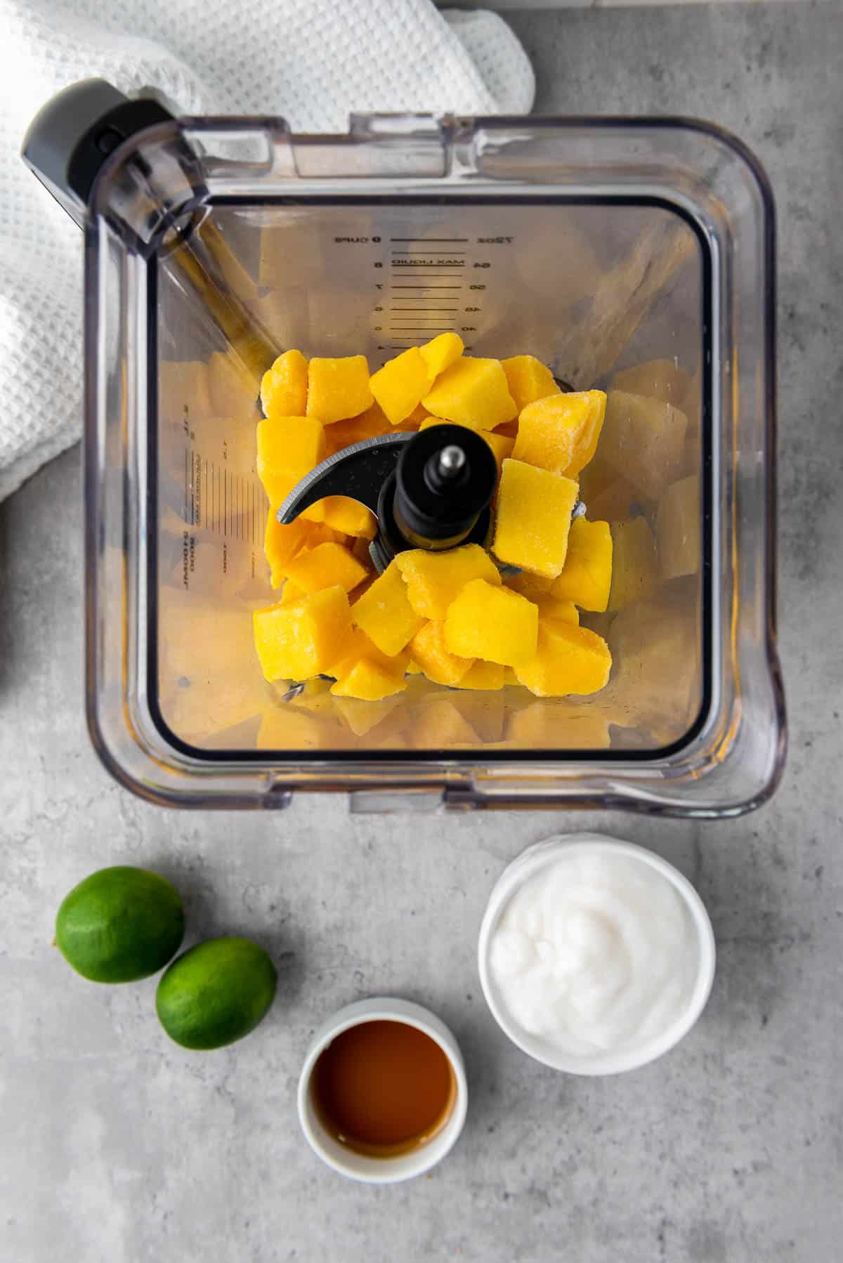 Chunks of mango in a blender with limes, yogurt, and maple syrup on the side, on top of a gray surface with a white napkin on the side.