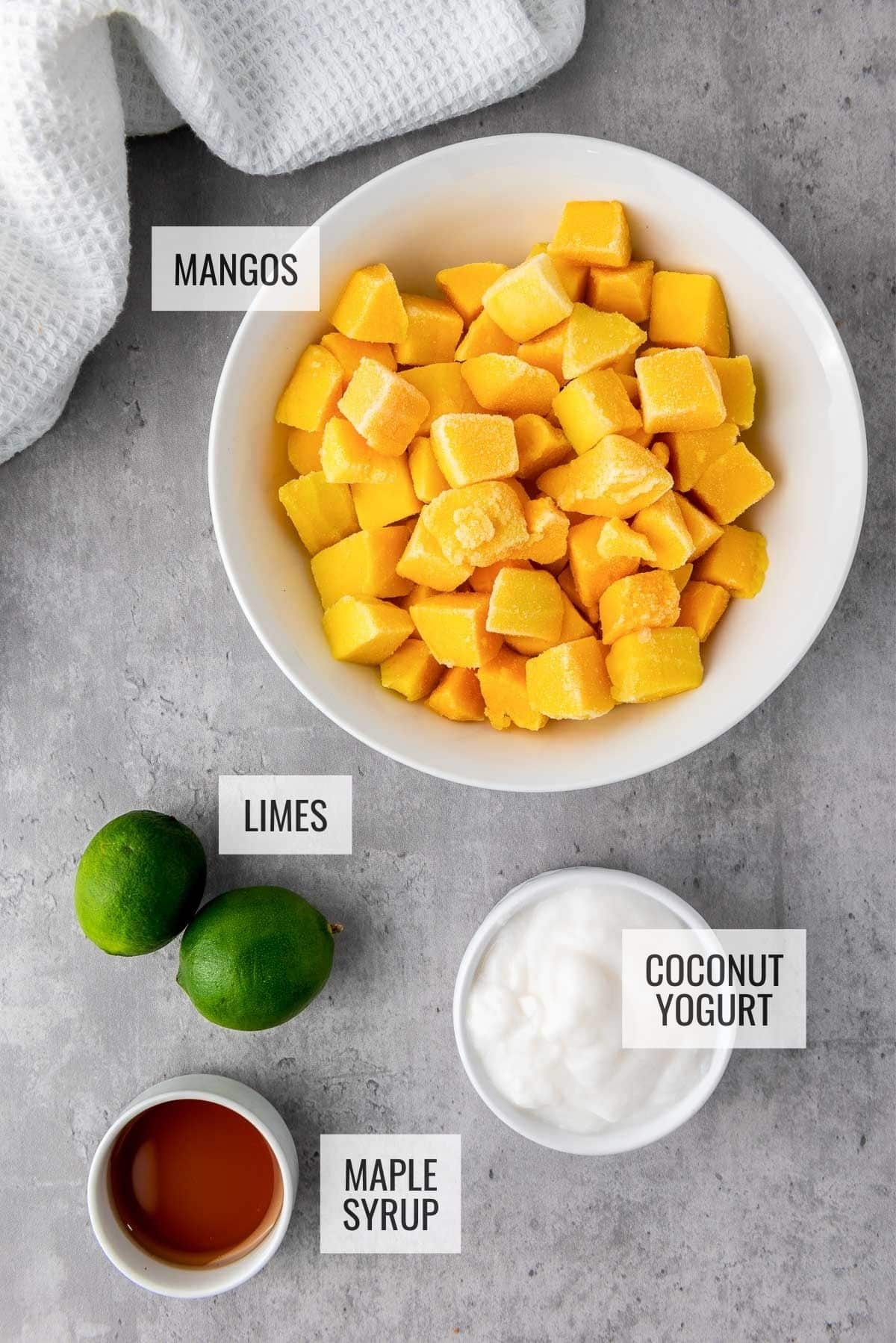 Labeled ingredients in bowls on top of a gray surface for making mango coconut lime popsicles, with a white napkin on the side.