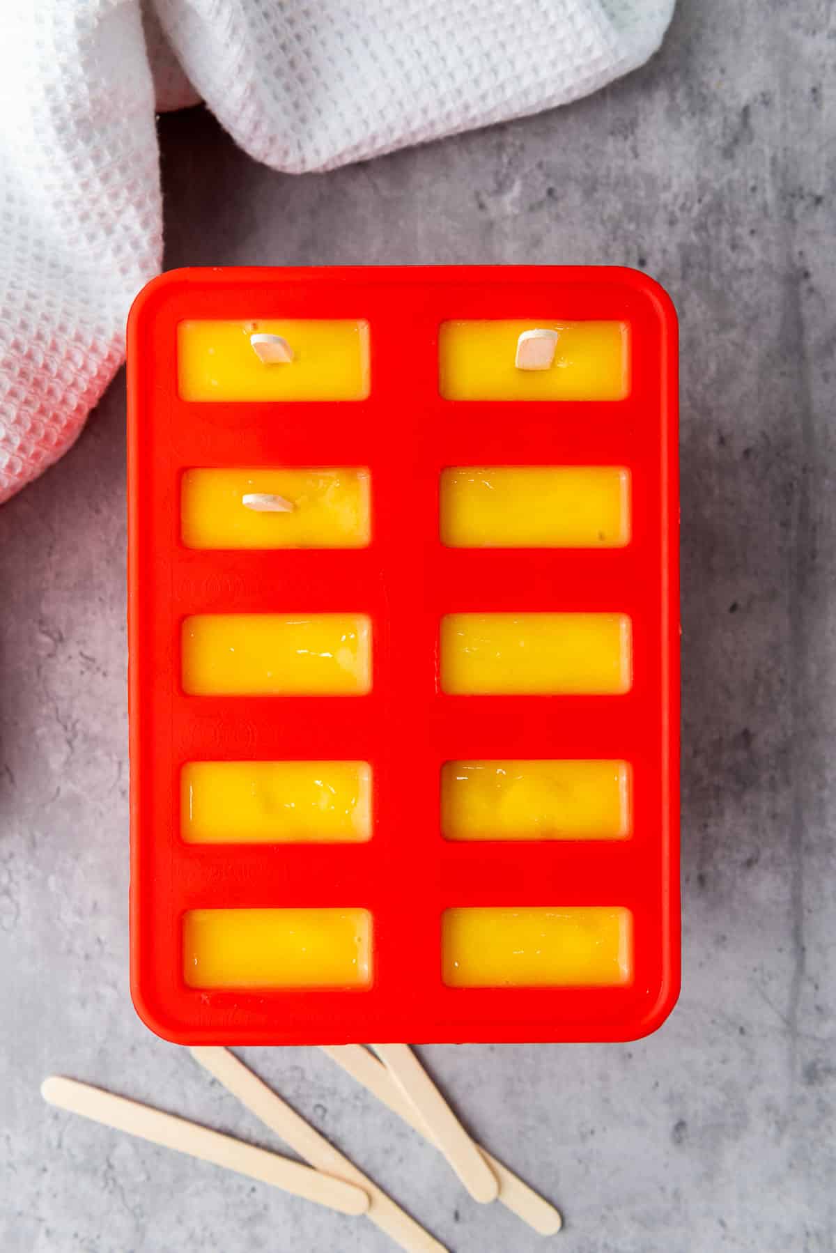 Mango coconut lime popsicles in a red popsicle mold ready for the freezer, on top of a gray surface with a white napkin on the side.