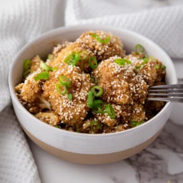 A fork inserted into a white bowl filled with Air Fryer Teriyaki Cauliflower Wings topped with sesame seeds and sliced scallions on top of a marble surface with a white napkin.