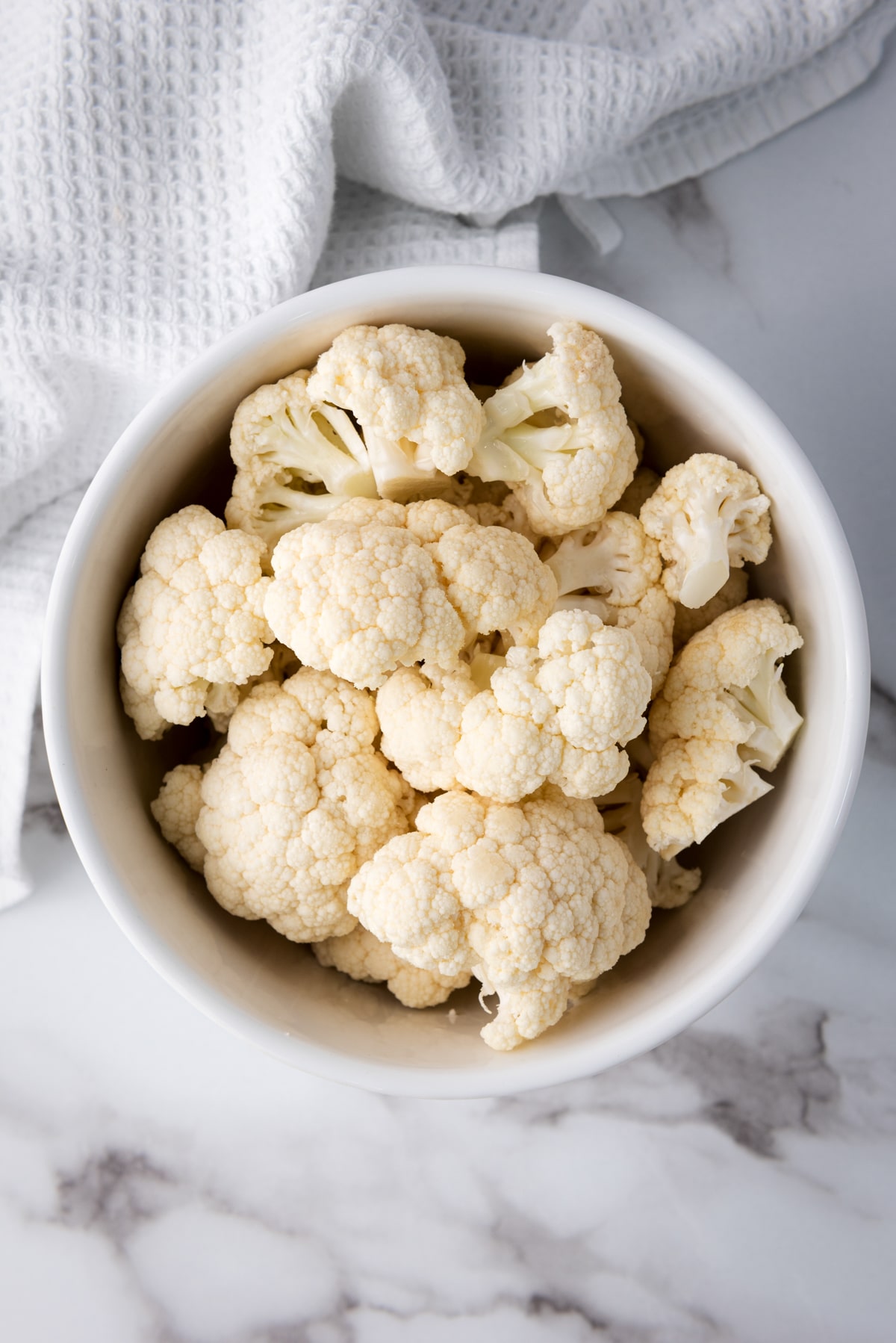 Raw cauliflower florets in a white bowl on top of a marble surface with a white napkin.