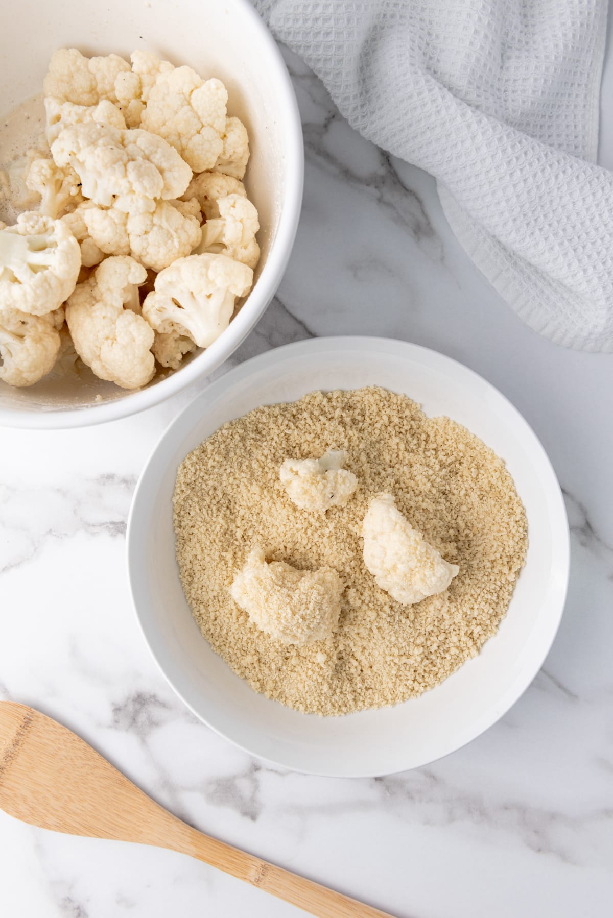 Cauliflower florets being dredged in white bowl of Panko bread crumbs with unbreaded cauliflower florets in a white bowl on the side, on top of a marble surface with a white napkin and a wooden spoon.