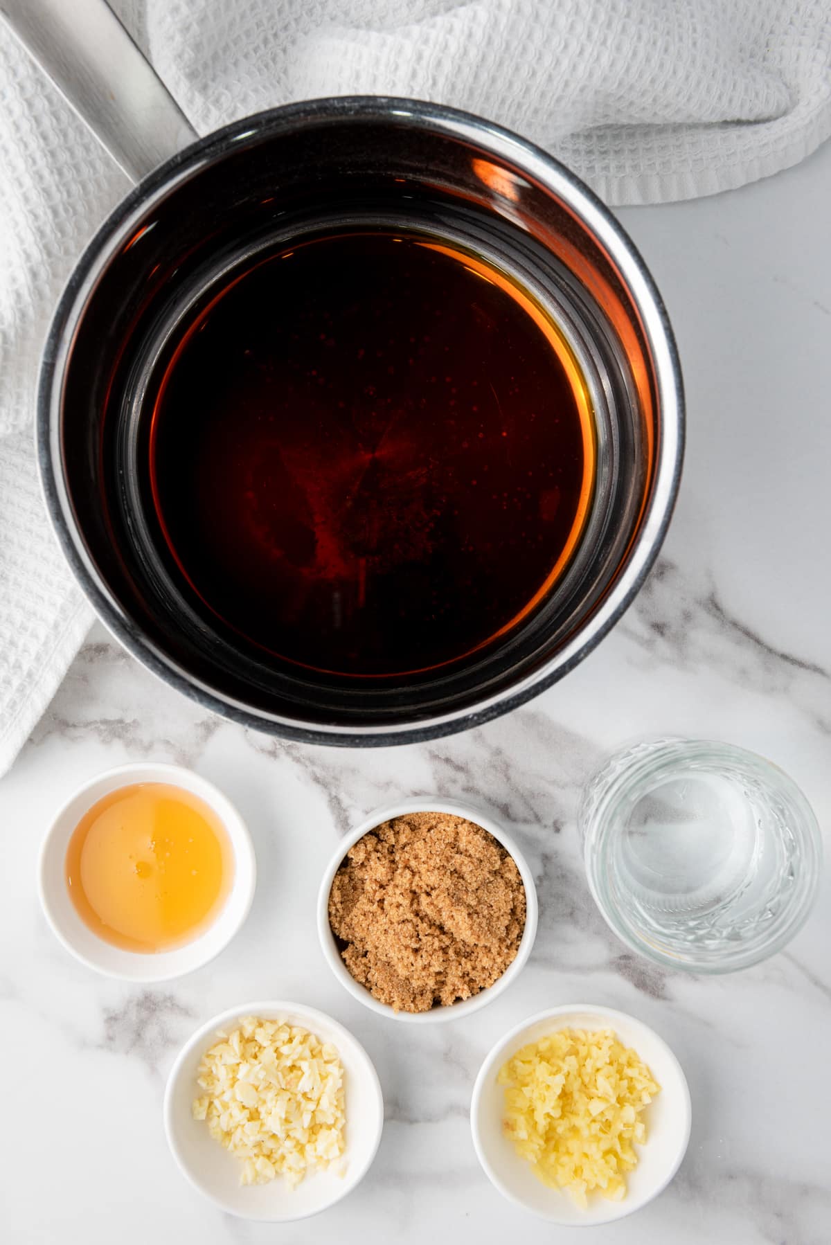 A small saucepan with soy sauce and other ingredients in small white bowls on the side on top of a marble surface with a white napkin.