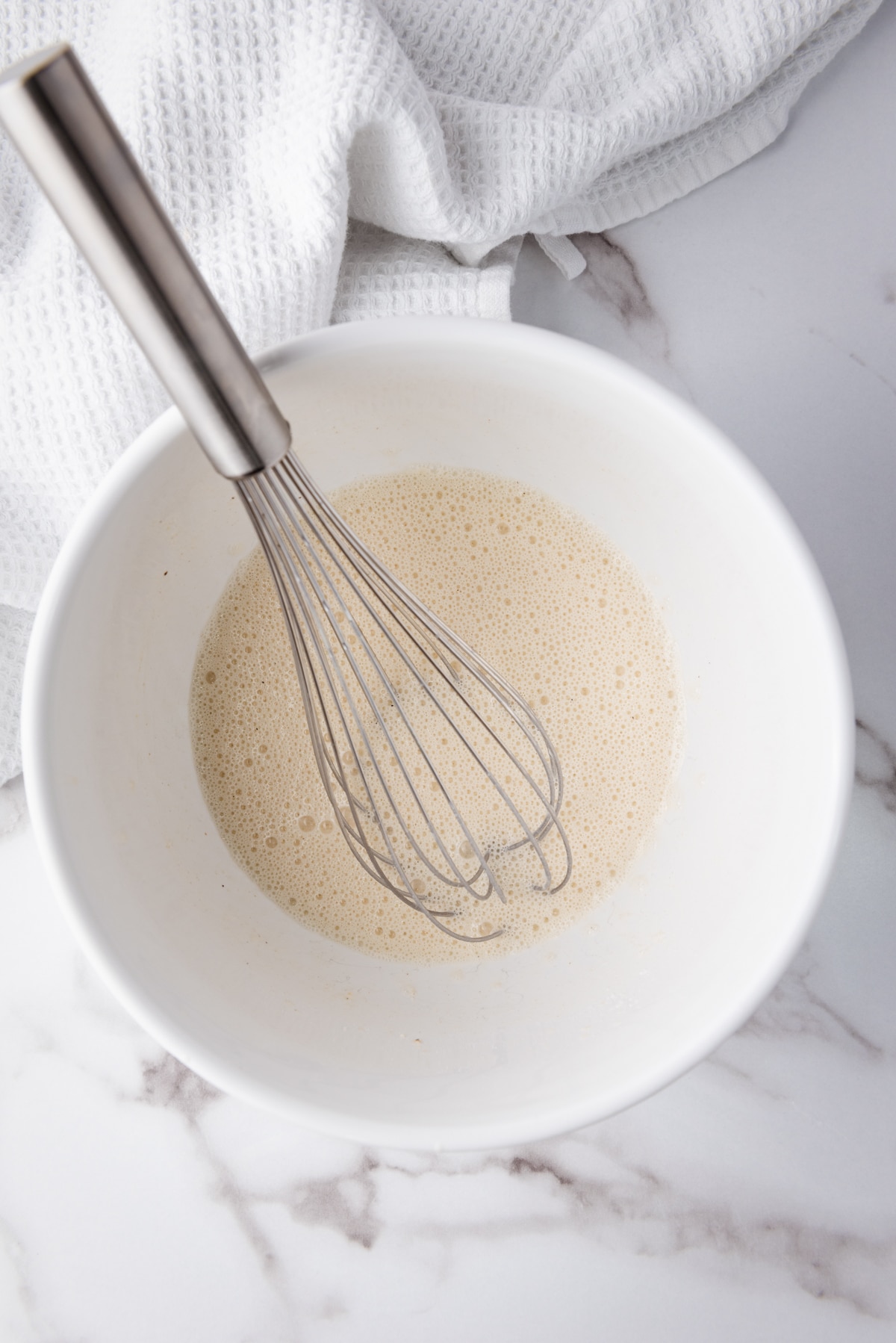 A whisk inserted into white bowl filled with milk on top of a marble surface with a white napkin.