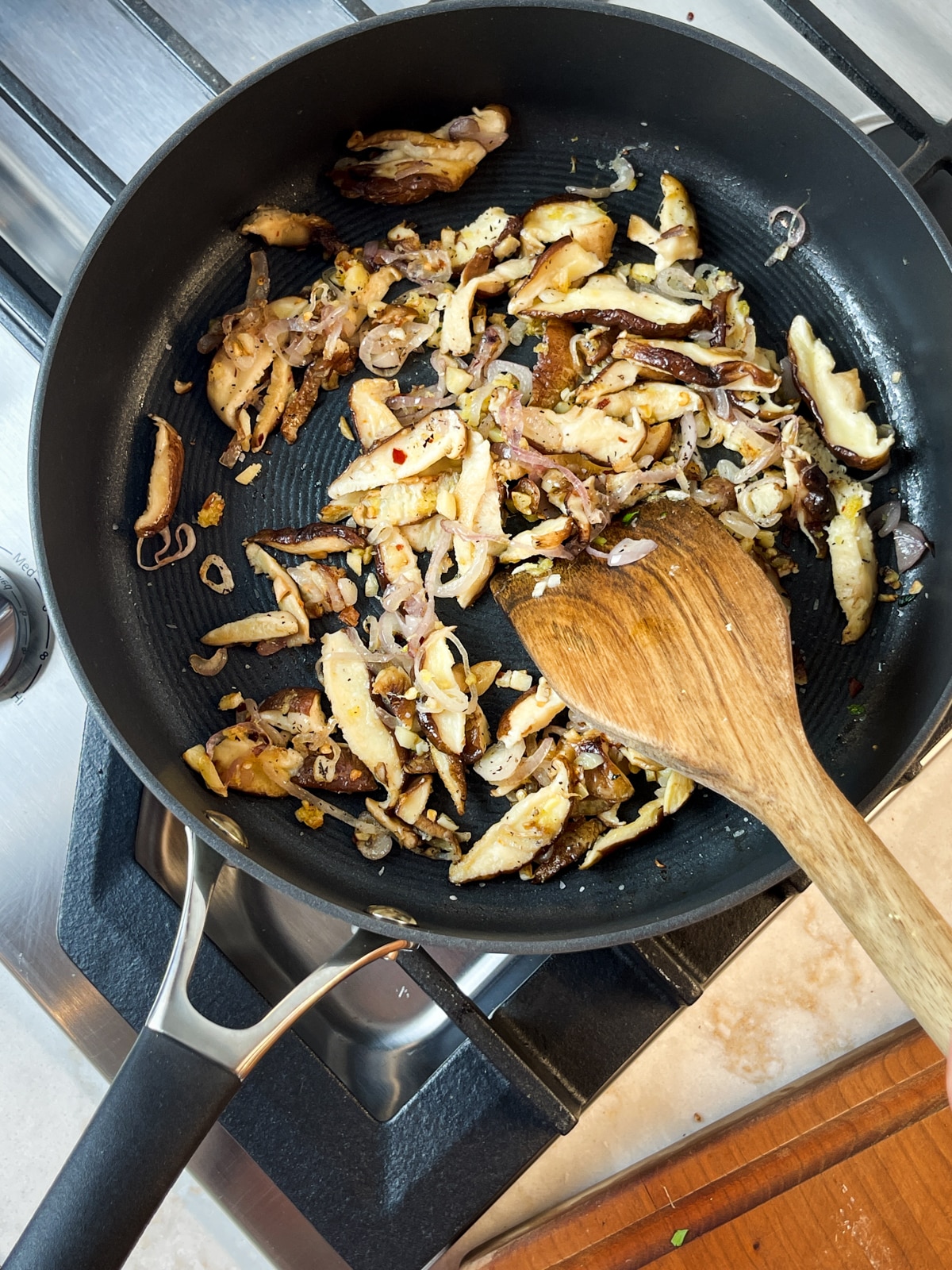 A wooden spatula stirring sliced shiitake mushrooms, shallots and garlic in a frying pan on a stove.