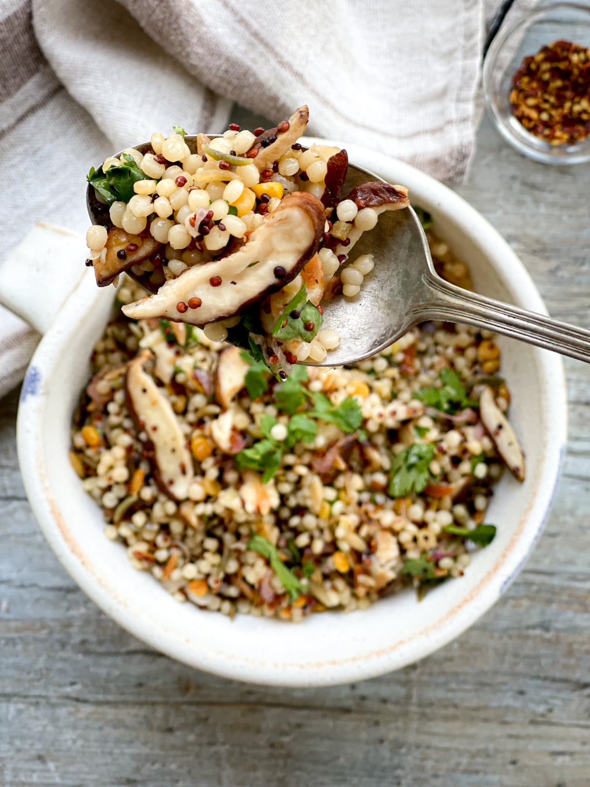 A spoonful of couscous and quinoa with shiitake mushrooms being held up above a white bowl on top a wooden board with a linen napkin and red pepper flakes on the side.