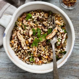 Couscous and quinoa with shiitake mushrooms in a white bowl with a fork on top of a wooden board with a linen napkin and red pepper flakes on the side.