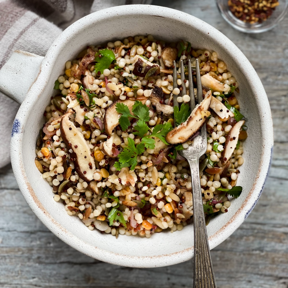 Couscous and quinoa with shiitake mushrooms in a white bowl with a fork on top of a wooden board with a linen napkin and red pepper flakes on the side.