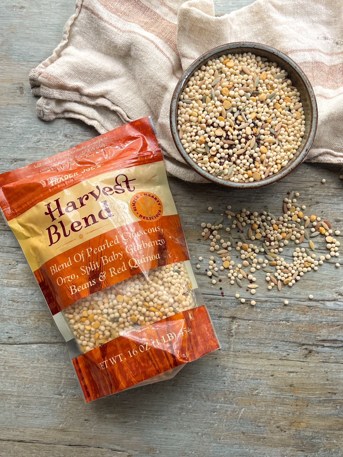 Trader Joe's Harvest Blend couscous and quinoa in a bag and in a bowl on the side on top of a wooden board with a linen napkin.