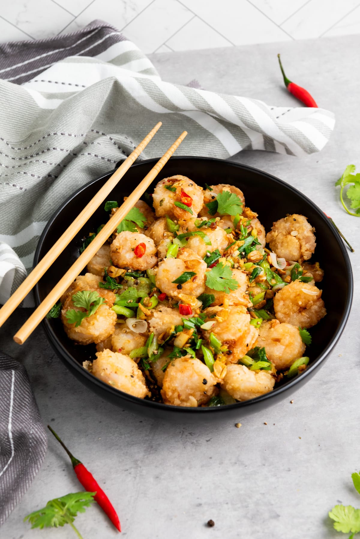 Chinese salt and pepper shrimp in a black bowl with sautéed vegetables and herbs, and a pair of chopsticks on top on a gray surface with a gray linen on the side.