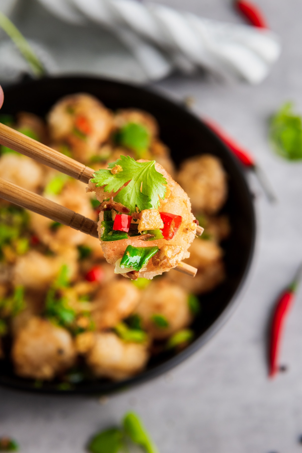A pair of chopsticks holding up a piece of Chinese salt and pepper shrimp above a black bowl filled with Chinese salt and pepper shrimp on top of a gray surface.