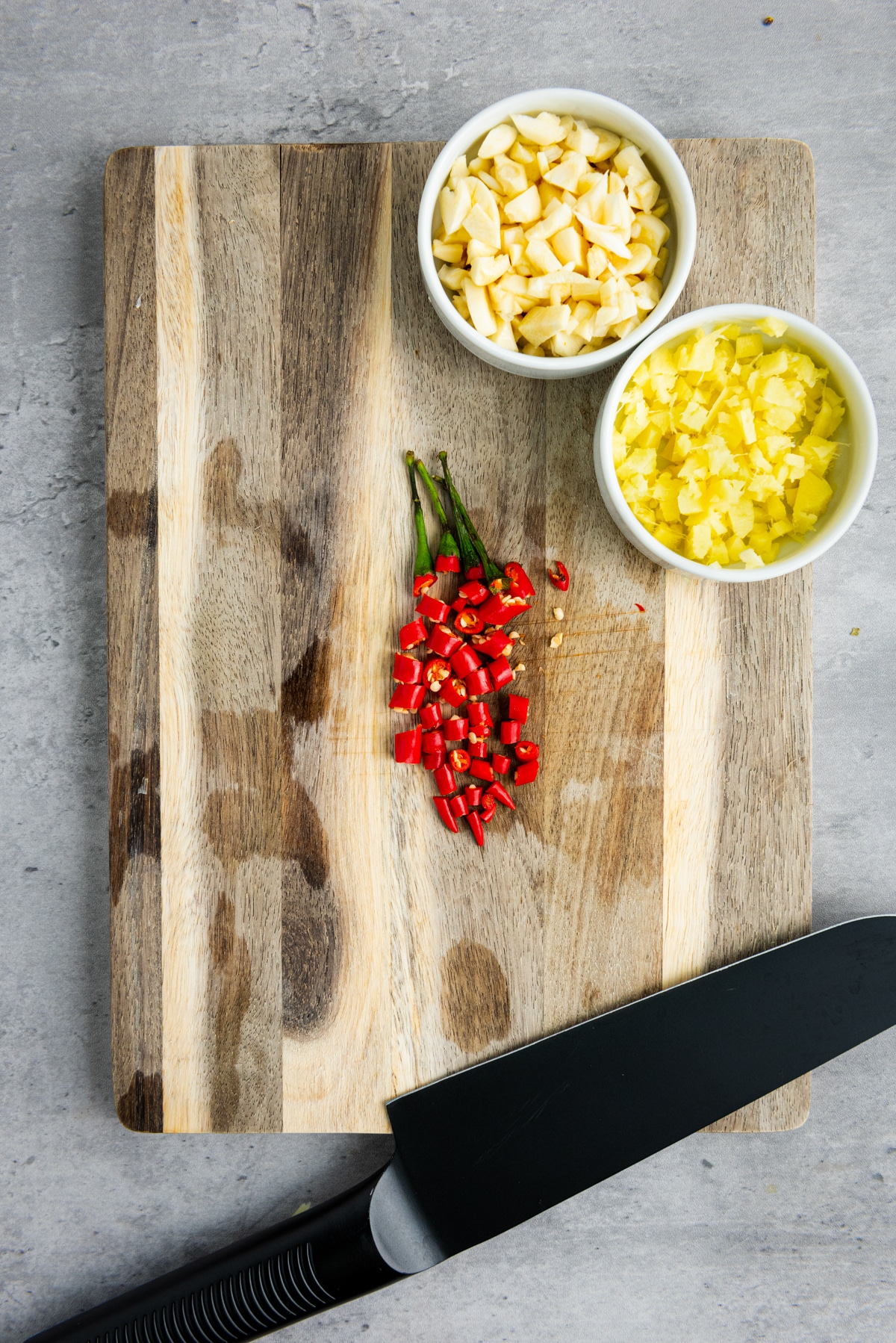 Chopped ginger and garlic in two small white bowls, and sliced red Thai chili peppers, on top of a wooden cutting board with a chef's knife on the side placed on top of a gray surface.