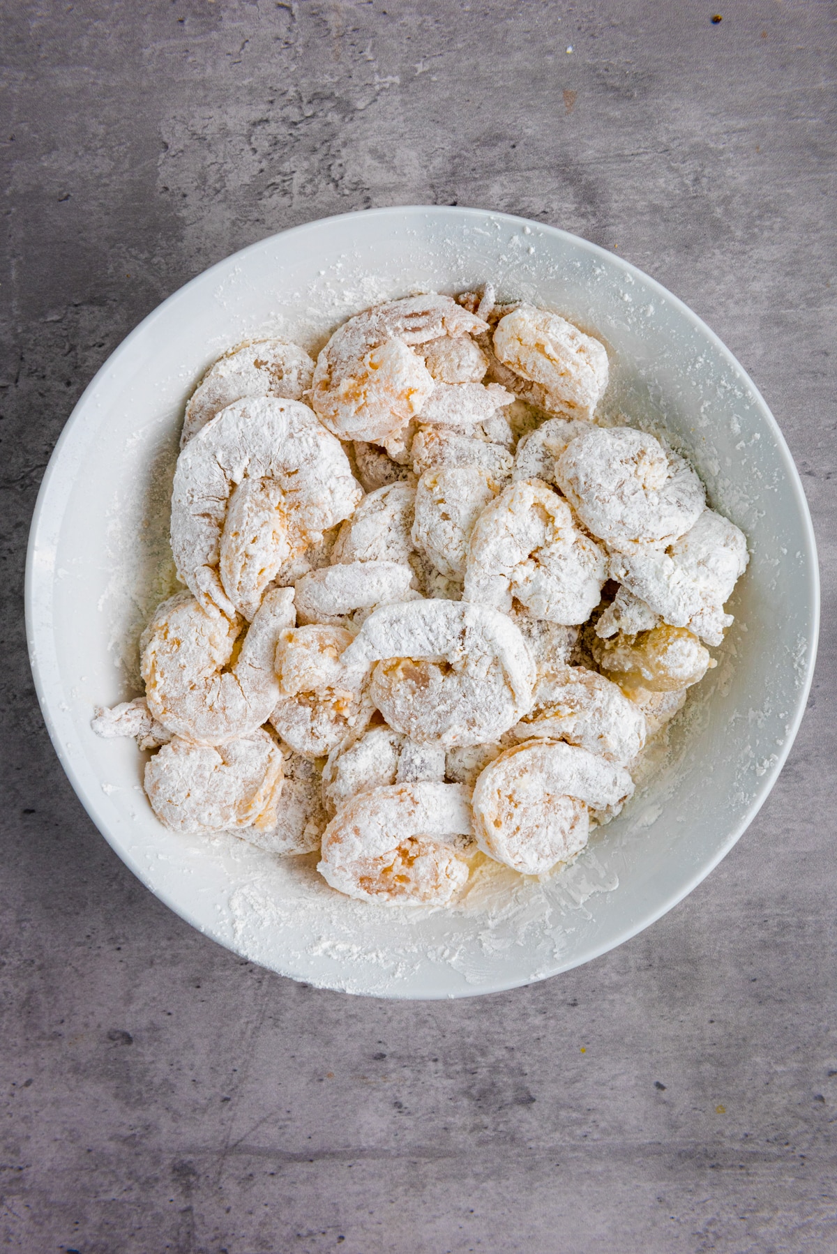 A white bowl filled with coated and dredged raw shrimp on top of a gray surface.