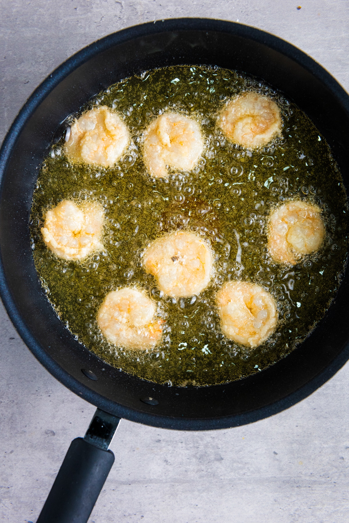 Shrimp being fried in oil in a frying pan on top of a gray surface.
