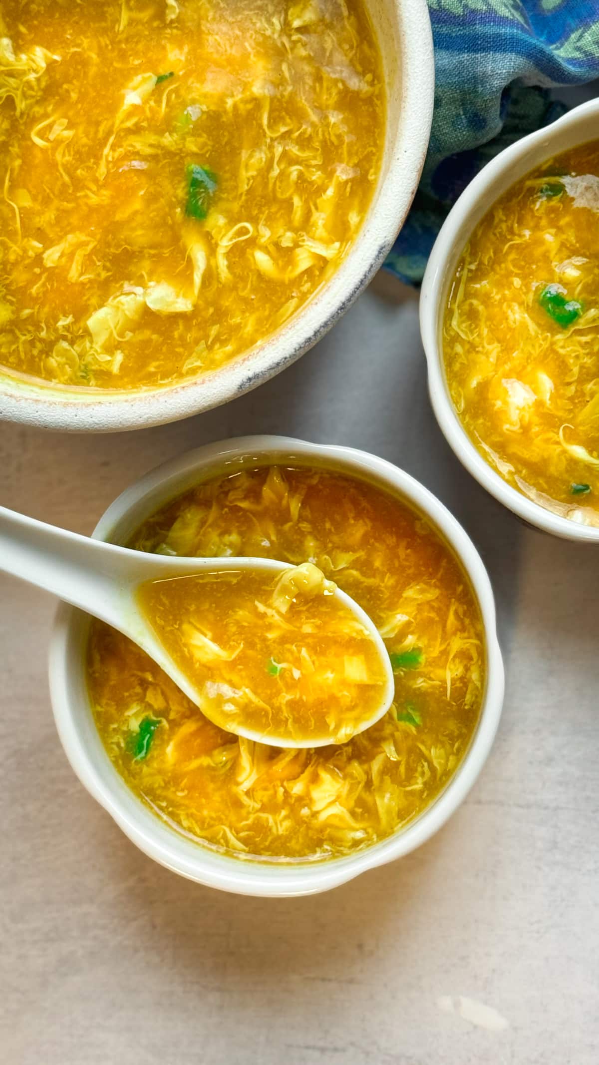 Chinese egg drop soup in three bowls with a white spoon inserted into the front bowl on top of a gray surface with a blue napkin on the side.