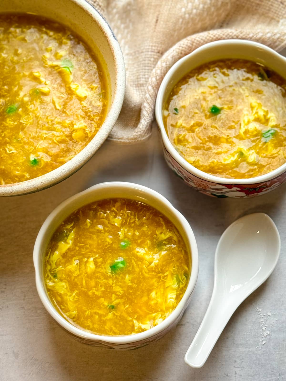 Chinese egg drop soup in three bowls with a white spoon on the side on top of a gray surface with a beige napkin on the side.