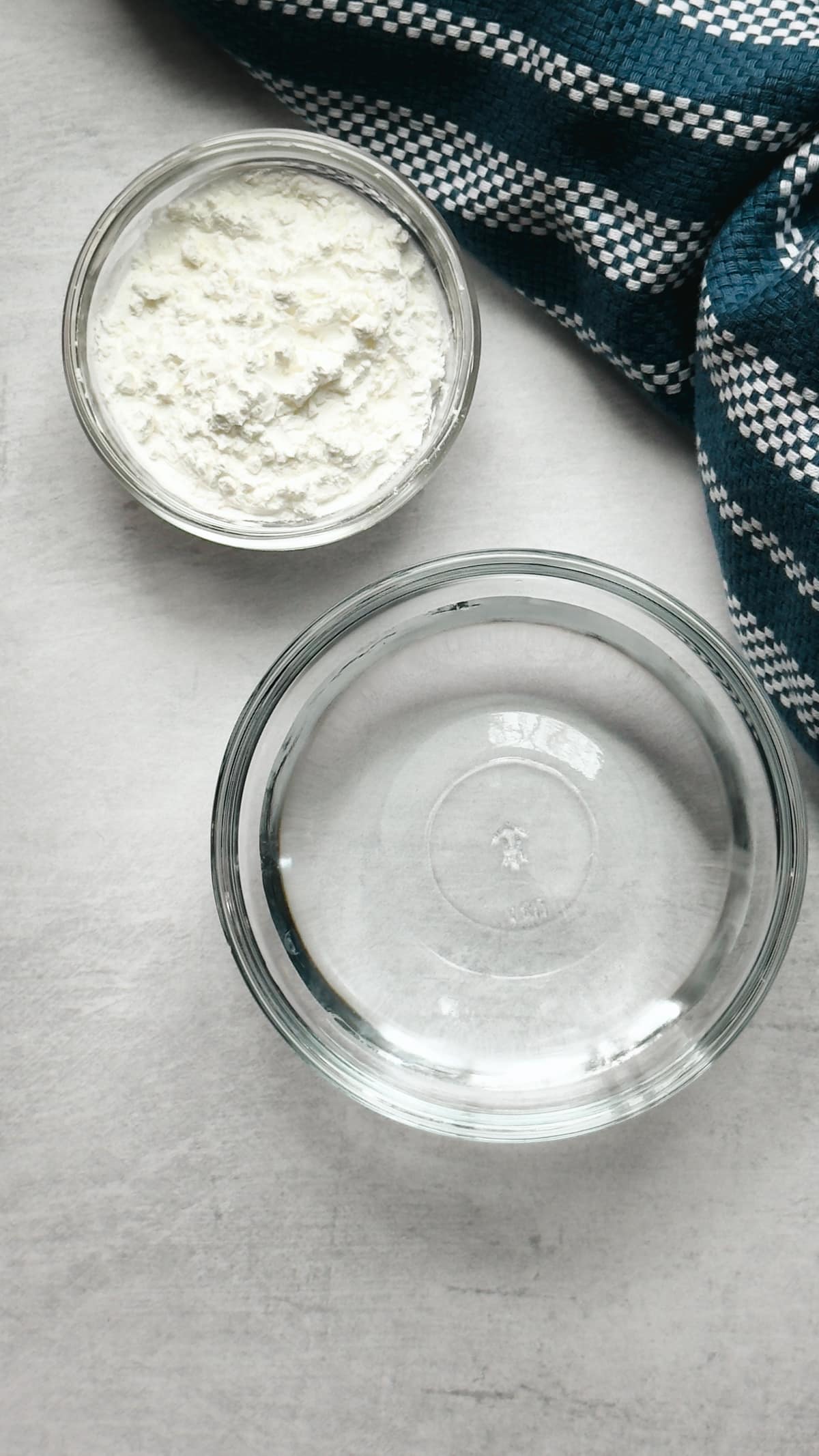 A glass bowl filled with water and a glass bowl with cornstarch on top of a gray surface with a blue napkin on the side.