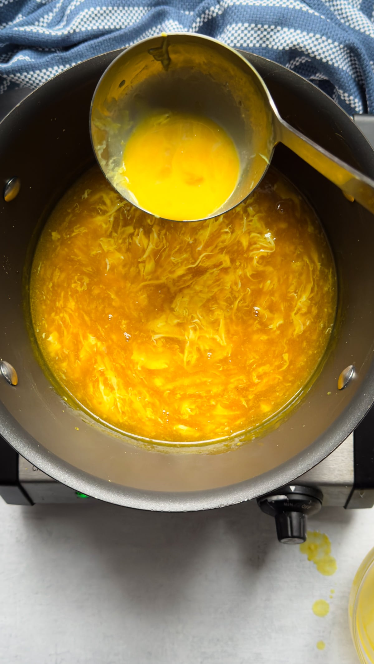 A silver ladle pouring eggs into a broth of Chinese egg drop soup in a pot on top of a gray surface with a blue napkin on the side.