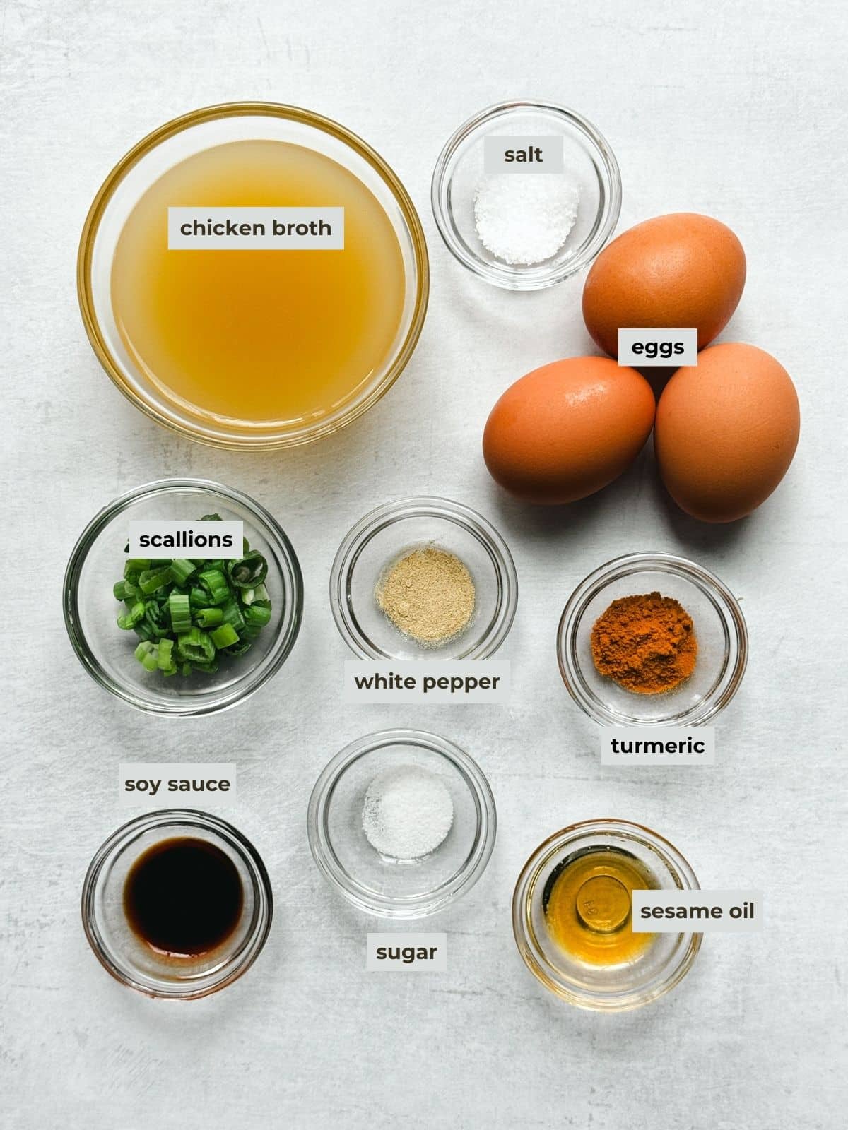 Labeled ingredients in glass bowls for making Chinese egg drop soup on top of a gray surface.