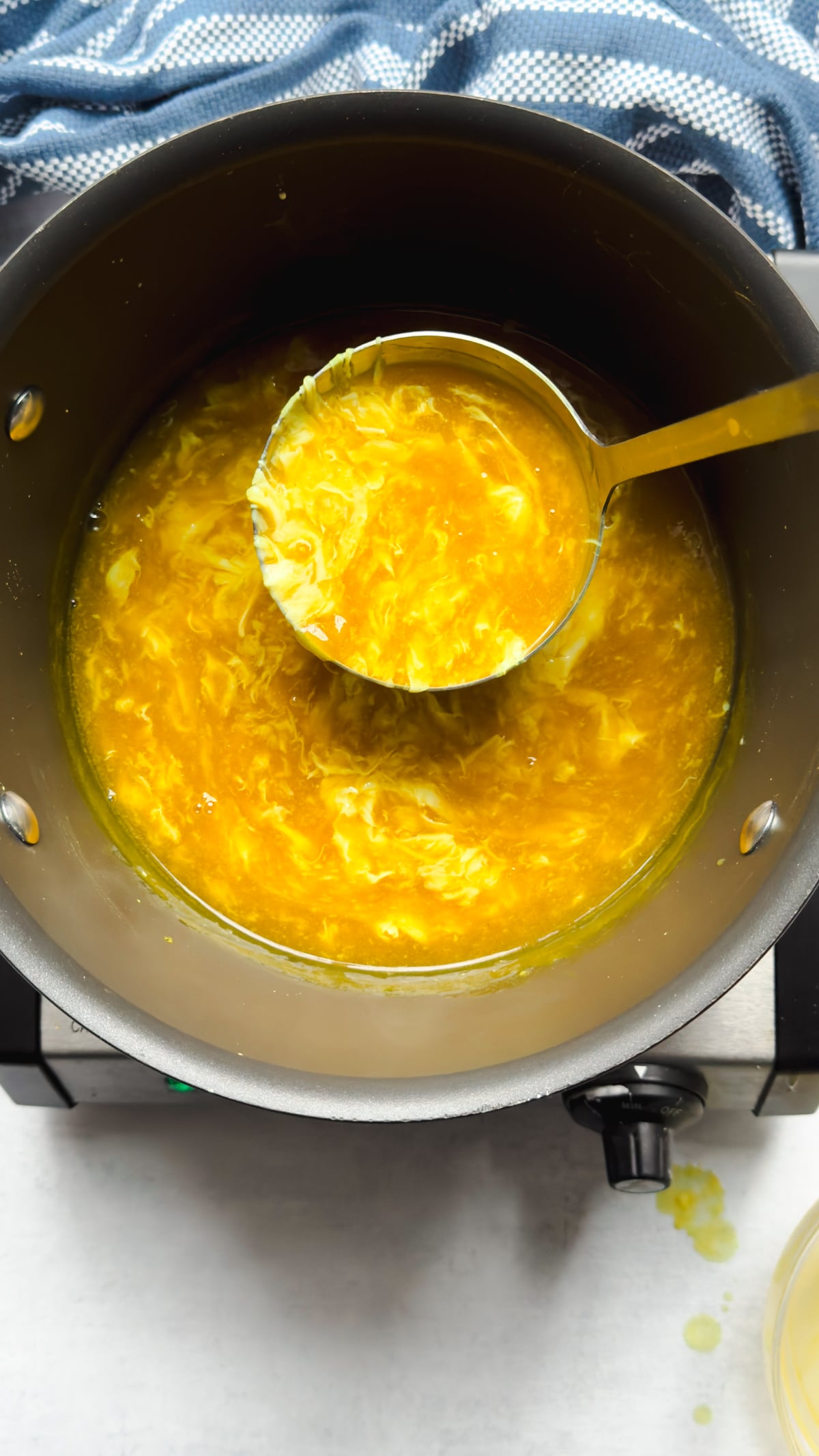 A silver ladle with a spoonful of Chinese egg drop soup being held above a pot of the soup on top of a gray surface with a blue napkin on the side.