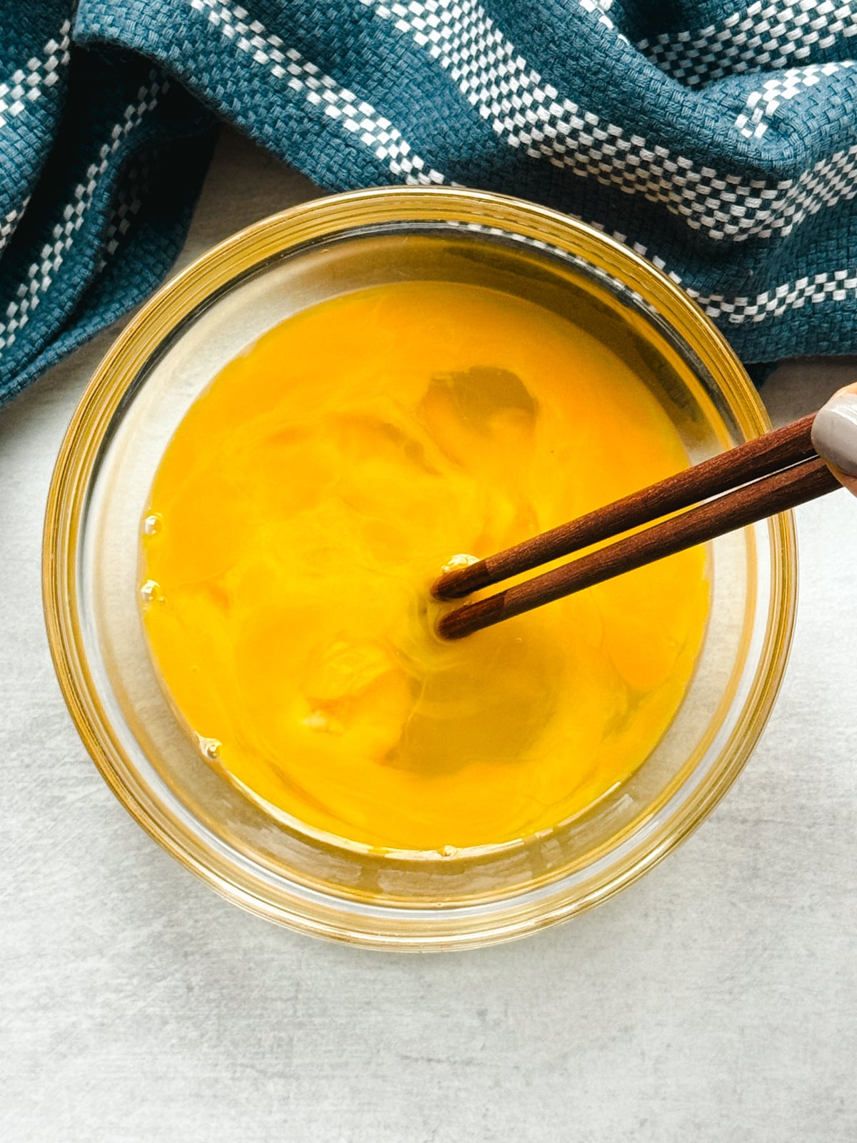 Eggs in a glass bowl being scrambled with chopsticks on top of a gray surface with a blue napkin on the side.
