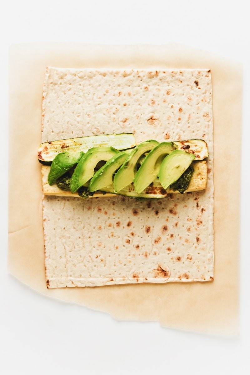 Assembling a grilled tofu and zucchini wrap on a white surface.