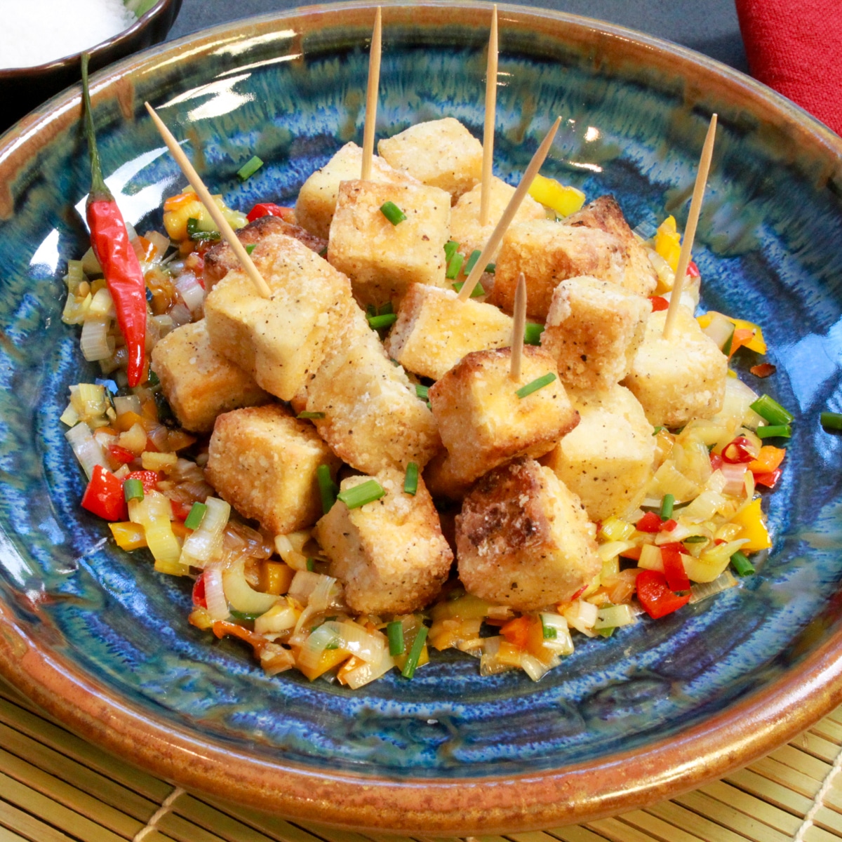 Crispy salt and pepper tofu cubes piled on top of sauteed vegetables on a round blue plate with a small bowl of salt on the side.