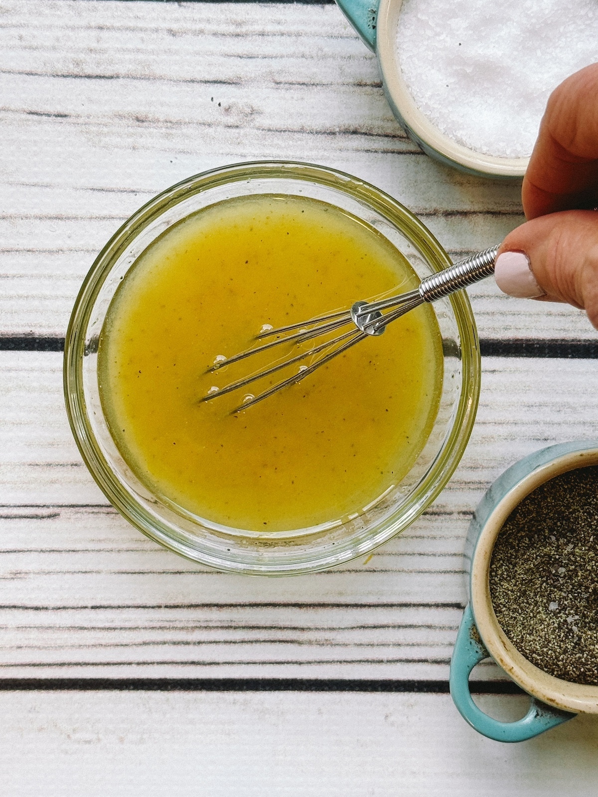 Yuzu lemon dressing in a clear bowl with a hand inserting a small whisk, and bowls of salt and pepper on the side, on top of a white wooden board.