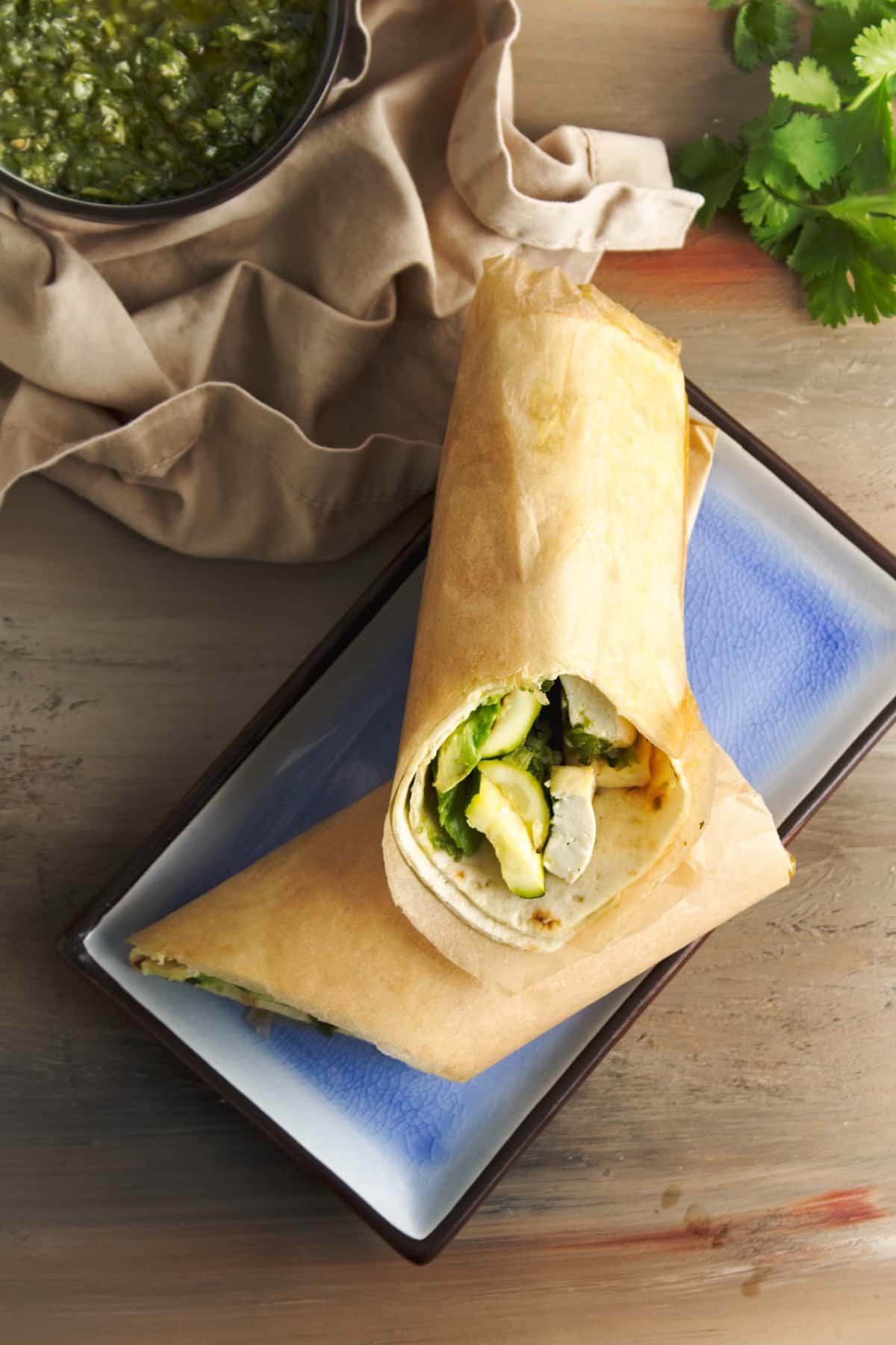 A grilled tofu and zucchini wrap sliced in half stacked on a blue plate with a napkin and sauce on the side, on top of a wooden board.