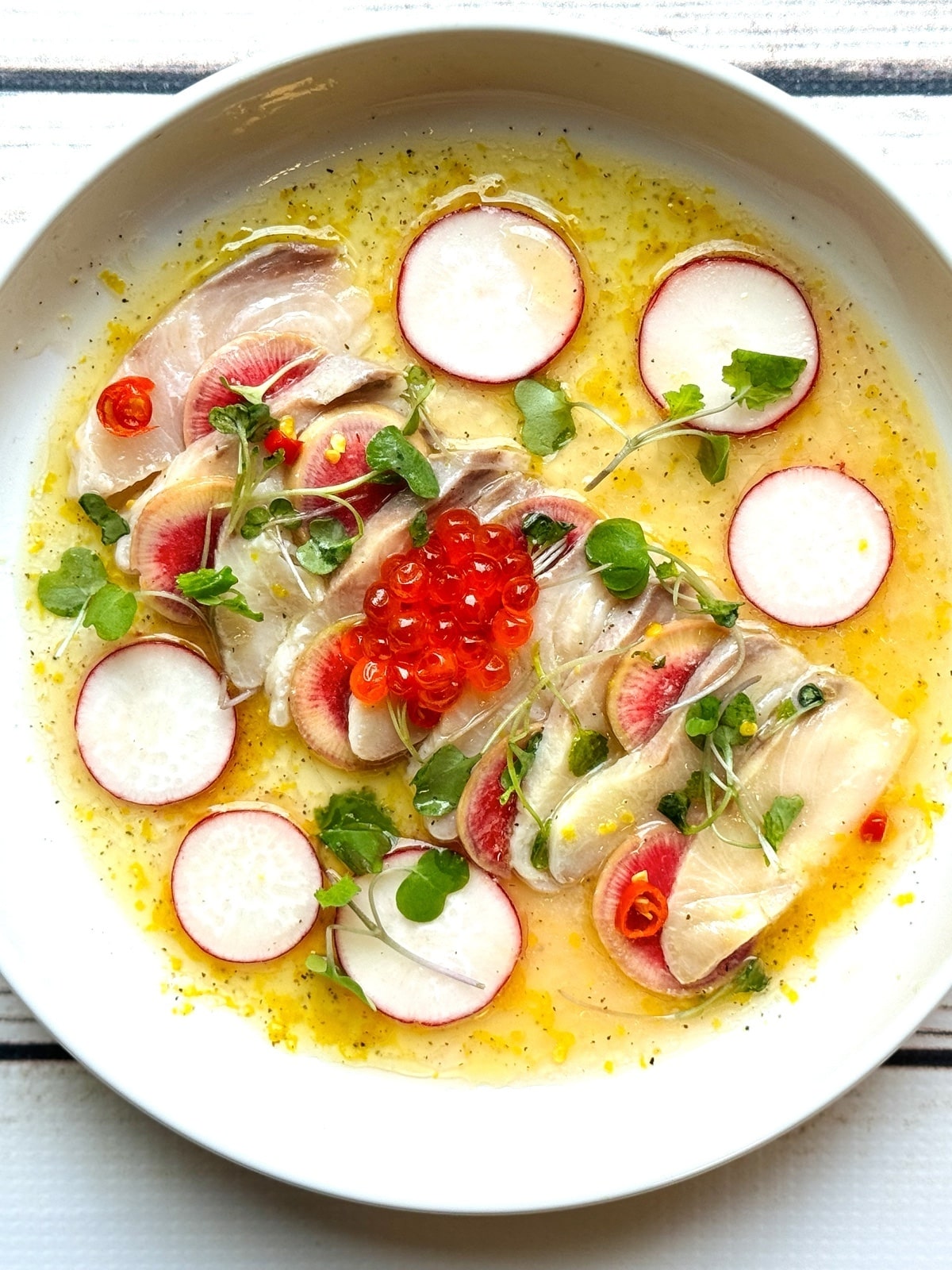 Hamachi yellowtail crudo on a white round plate with a yuzu lemon dressing surrounded by shaved radishes and topped with red fish eggs and microgreens.