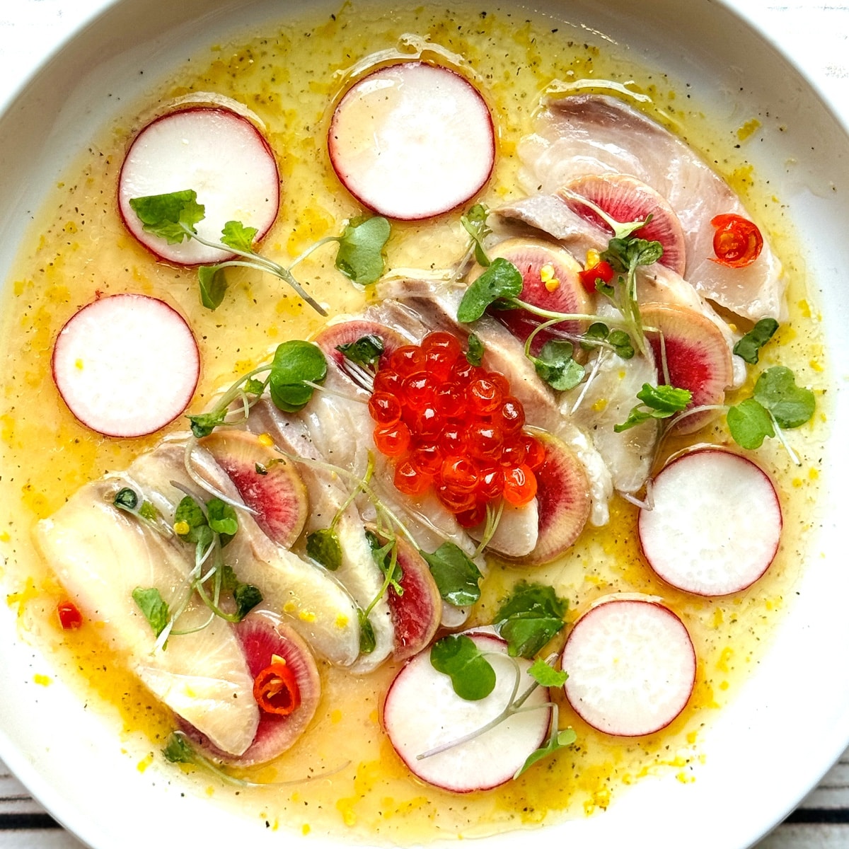 Hamachi yellowtail crudo on a white round plate with a yuzu lemon dressing surrounded by shaved radishes and topped with red fish eggs and microgreens.