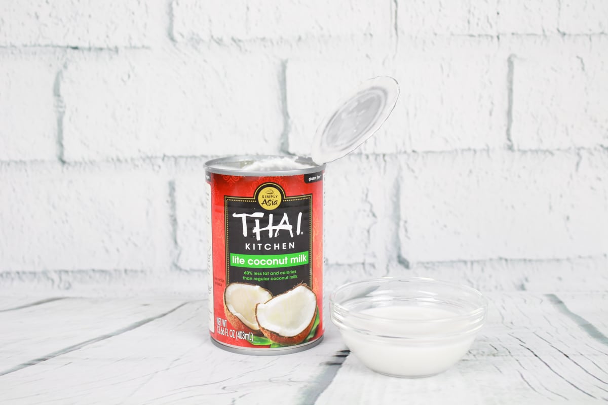 A can of Thai Kitchen coconut milk and a small glass bowl filled with coconut milk placed on top of a white plank board with white brick in the background.