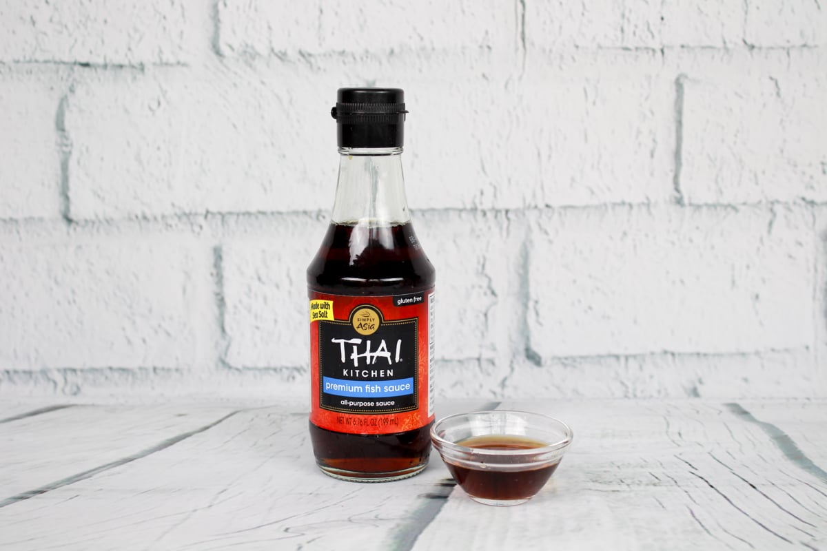 A bottle of Thai Kitchen fish sauce and a small glass bowl filled with fish sauce placed on top of a white plank board with white brick in the background.