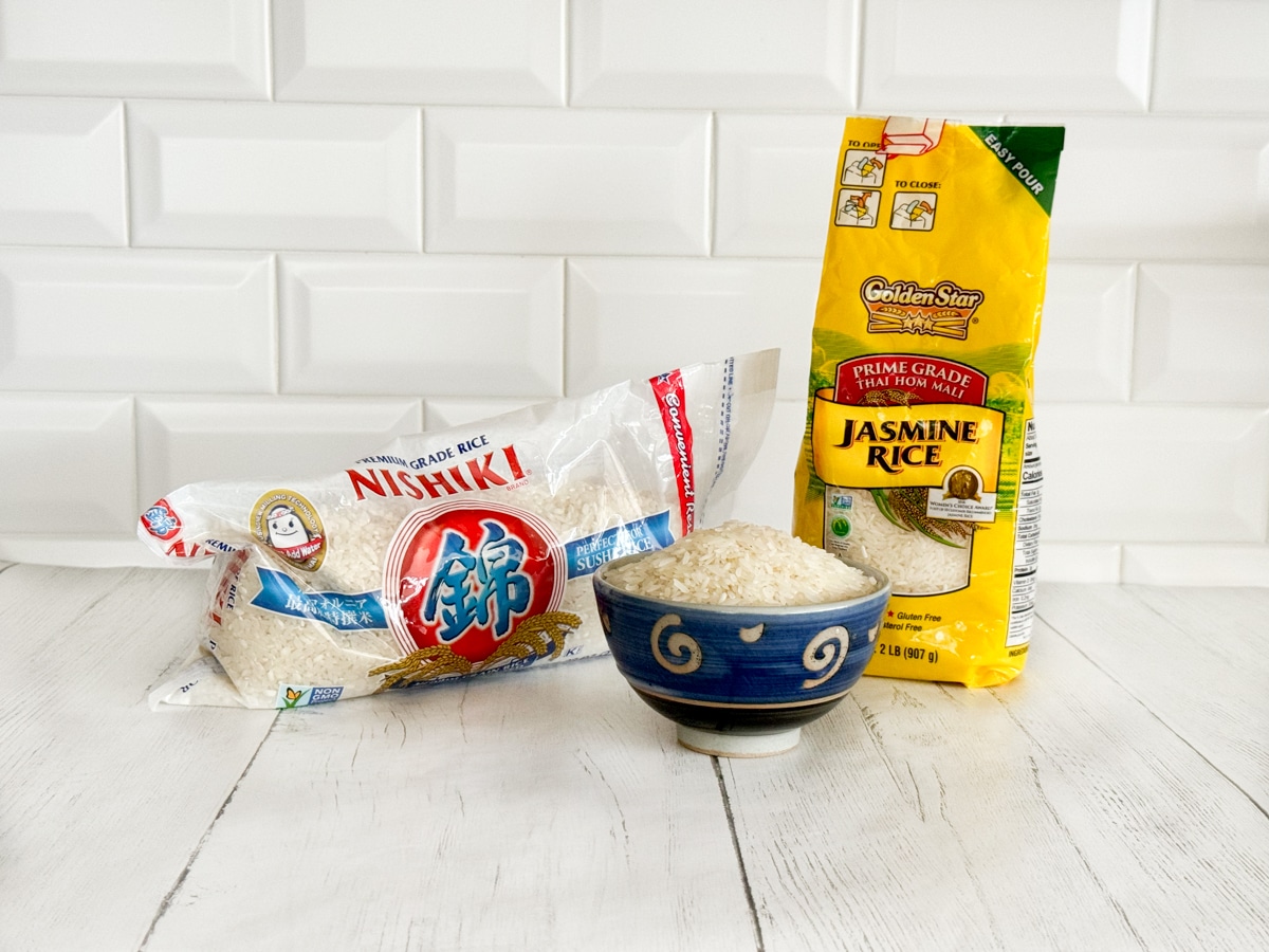 A bag of sushi rice and a bag of Jasmine rice, and a blue bowl of raw rice, on top of a white plank board with a white brick background.