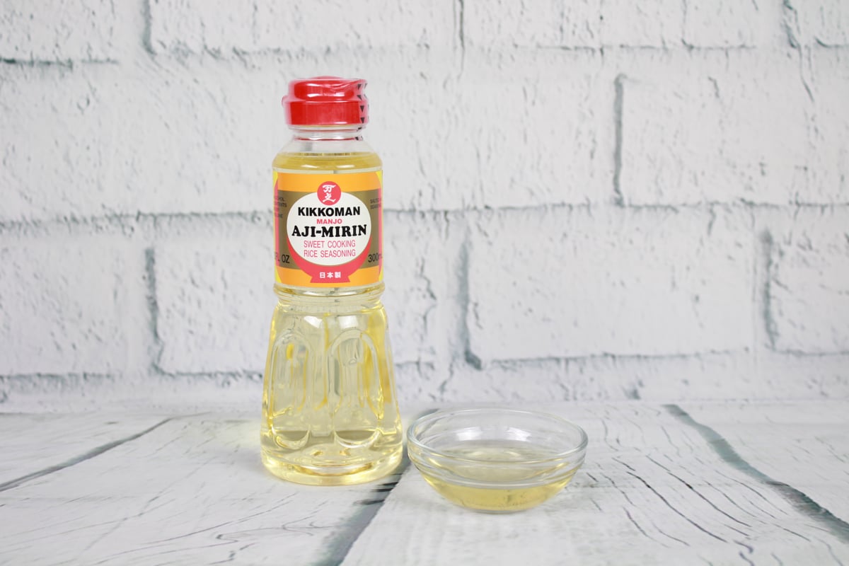 A bottle of Kikkoman mirin and a small glass bowl filled with mirin placed on top of a white plank board with white brick in the background.