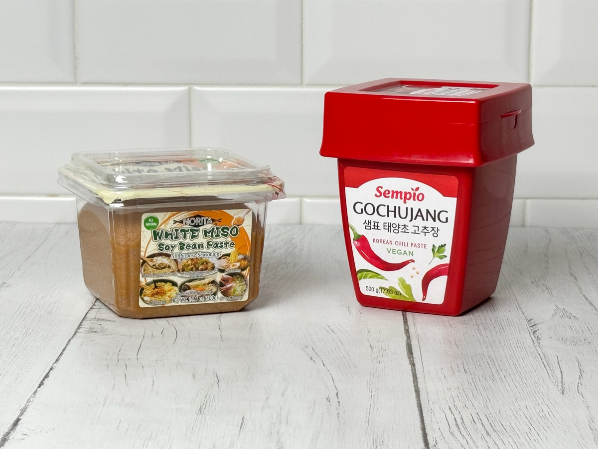 A container of miso paste and a container of gochujang paste on top of a white plank board with a white brick background.